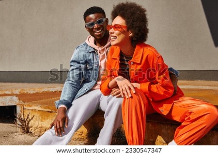 Cheerful young african american best friends in trendy outfits and sunglasses laughing and having fun while sitting on rusty stairs near building on urban street, trendy friends in urban settings Royalty-Free Stock Photo #2330547347