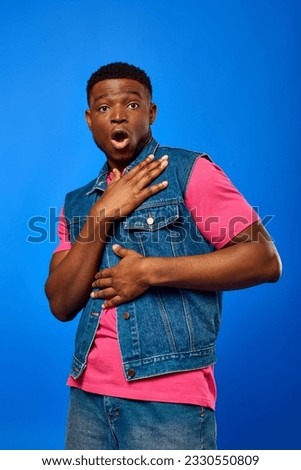 Shocked young african american man with modern hairstyle wearing denim vest and bright t-shirt while looking at camera and standing isolated on blue, trendy man showing summer style Royalty-Free Stock Photo #2330550809