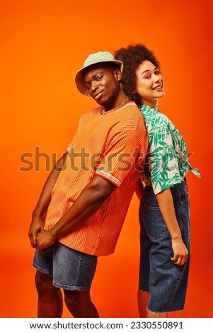 Young african american man in summer outfit and panama hat looking at camera and standing back to back with best friend with bold makeup and posing isolated on orange, friends in trendy clothes