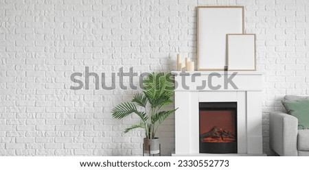 Electric fireplace with blank frames and houseplant near white brick wall in living room. Banner for design