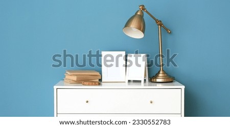 Blank frames, lamp and books on chest of drawers near blue wall. Banner for design