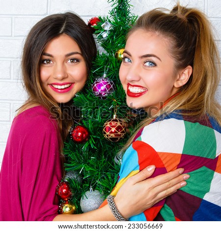 Close up lifestyle indoor portrait of two young woman posing near decorated Christmas tree, at New Year eve. Smiling having fun, ready for celebration. Bright holiday image of best friends.