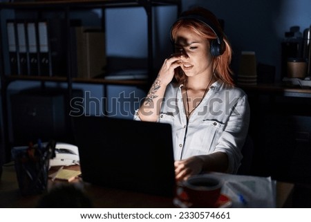 Young caucasian woman working at the office at night smelling something stinky and disgusting, intolerable smell, holding breath with fingers on nose. bad smell  Royalty-Free Stock Photo #2330574661