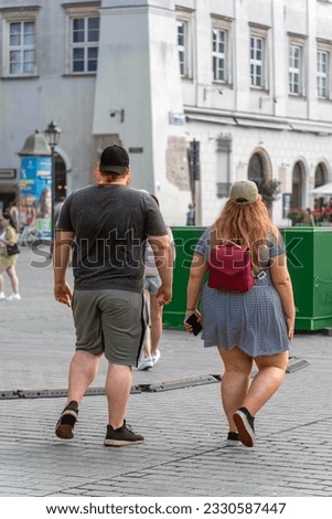 Overweight couple, backpack and smartphone. Plump couple in trendy attire walking with confidence on city sidewalk. Blurred background of busy street, diverse passers-by. Body-positive representation