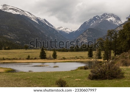 Green nature setting with a pond in foreground and a pale, cloudy sky.  Rocky Mountain pond with reflective water, green grass and dark mountains.  Colorado high country with cloudy sky. Royalty-Free Stock Photo #2330607997