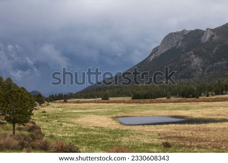 Green nature setting with a pond in foreground and a dark, cloudy sky.  Rocky Mountain pond with reflective water, green grass and dark mountains.  Colorado high country with cloudy sky. Royalty-Free Stock Photo #2330608343
