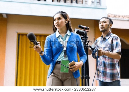 a beautiful reporter in blue cardigan with green name tag hunting for news with a cameraman behind her
