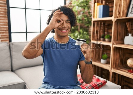 Hispanic man with curly hair holding credit card smiling happy doing ok sign with hand on eye looking through fingers 