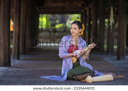 Beautiful pretty young Asian woman wearing a traditional Thai dress Lanna style sit with a garland flower and umbrella in Wat Ton Kwen Temple Chiang Mai, Thailand. Portrait the old fashion costume. Royalty-Free Stock Photo #2330644031