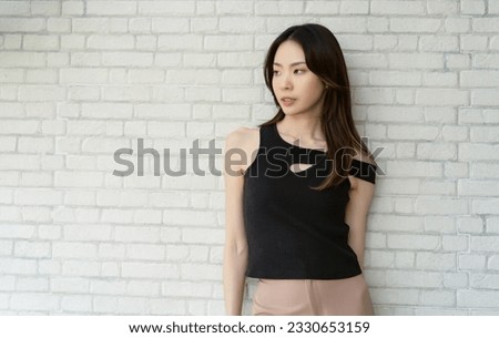 Positive serious millennial Asian woman wearing off shoulder black and looking away on white brick wall background. Cosmetic care, beautiful Korean or Japanese girl with confidence.