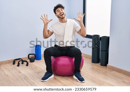 Hispanic man with beard sitting on pilate balls at yoga room celebrating mad and crazy for success with arms raised and closed eyes screaming excited. winner concept 