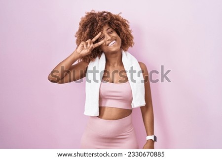 Young hispanic woman with curly hair wearing sportswear and towel doing peace symbol with fingers over face, smiling cheerful showing victory 