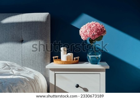 Stylish modern bedroom in dark colors. Cozy interior with navy blue walls, home decor. Bed with grey fabric headboard, white blanket, bedside table, vase with pink hydrangea flower, candles on tray. Royalty-Free Stock Photo #2330673189