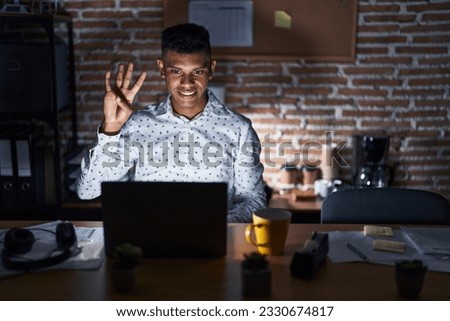 Young hispanic man working at the office at night showing and pointing up with fingers number four while smiling confident and happy. 