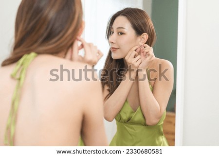 Get dress, elegant asian young woman with makeup glamour, getting ready wearing green cloth, gorgeous female putting earring or jewelry looking into the mirror at home, prepare going event night party Royalty-Free Stock Photo #2330680581