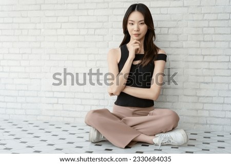 Portrait of beautiful young Asian woman wearing off shoulder top sitting on floor and looking at camera on white brick wall background. Millennials Korean or Japanese girl lifestyle.