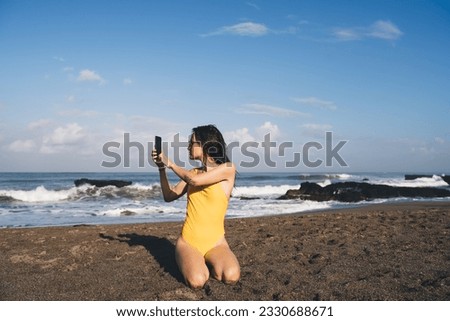 Side view of young woman in sunglasses while kneeling on sandy beach twisting spine to take pictures of sea waves with mobile phone and sitting on sandy beach against blue cloudy sky