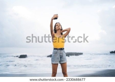 Positive young female looking up at screen of mobile phone taking selfie with hand touching back of neck while standing on seashore and enjoying summer vacation in tropical country