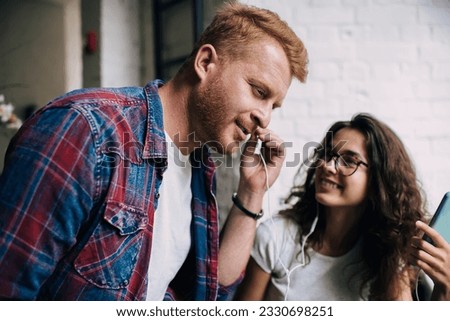 Positive young man smiling and looking away while listening to music shared by female friend in earphones attached to smartphone and enjoying weekend in cafe Royalty-Free Stock Photo #2330698251