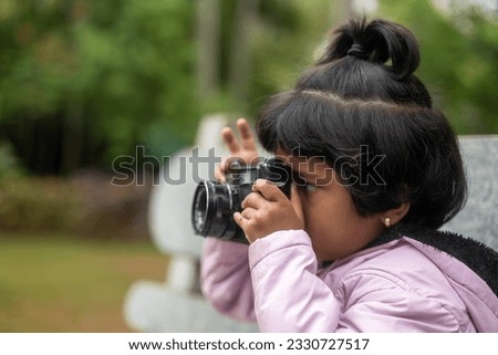 A young girl with pigtails and a big smile holds a DSLR camera up to her eye. She is looking through the viewfinder, focused on taking a picture.