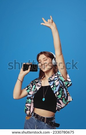 Vertical photo of happy young girl listening music with headphones and holding smartphone while showing rock sign, isolated on blue background. People, emotions and lifestyle concept.