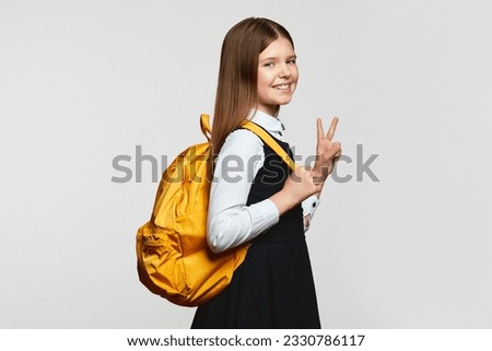 Excited schoolgirl with backpack smiling for camera and gesturing V sign while standing isolated over white background. Back to school concept