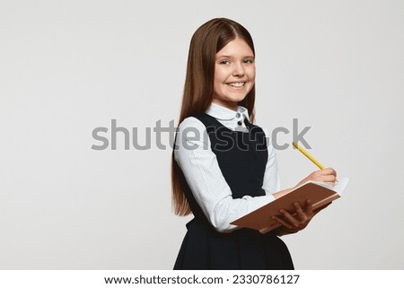 Cute nerdy preteen girl in school uniform smiling and looking at camera while writing in the notebook against white background with free space