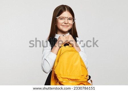 Cheerful nerdy school girl wearing uniform and eyeglasses holding shiny yellow backpack, smiling and looking at camera against white background