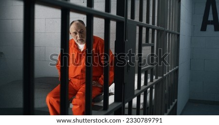 Elderly criminal in orange uniform sits on prison bed and thinks about freedom. Prisoner serves imprisonment term in jail cell. Guilty inmate in detention center or correctional facility. Royalty-Free Stock Photo #2330789791