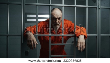 Elderly prisoner in orange uniform holds hands on metal bars, looks at camera. Criminal serves term of imprisonment in prison cell. Inmate stands behind bars in jail or detention center. Portrait. Royalty-Free Stock Photo #2330789835