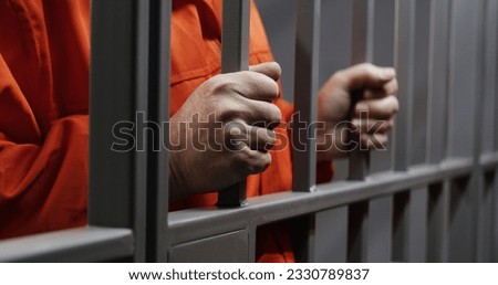 Hands close up of elderly prisoner in orange uniform holding metal bars, standing in prison cell. Criminal serves imprisonment term for crime. Inmate in jail or correctional facility. Justice system. Royalty-Free Stock Photo #2330789837