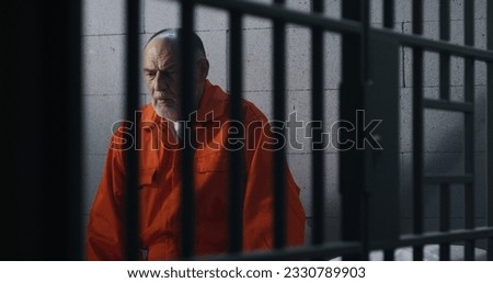 Elderly criminal in orange uniform sits on prison bed and thinks about freedom. Prisoner serves imprisonment term in jail cell. Guilty inmate in detention center or correctional facility. Royalty-Free Stock Photo #2330789903
