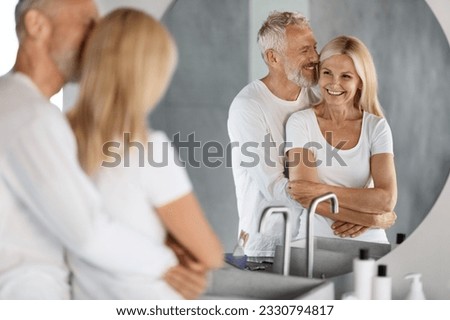 Happy Beautiful Senior Couple Looking At Mirror In Bathroom And Embracing, Romantic Mature Spouses Hugging While Getting Ready Together At Home In The Morning, Selective Focus On Reflection