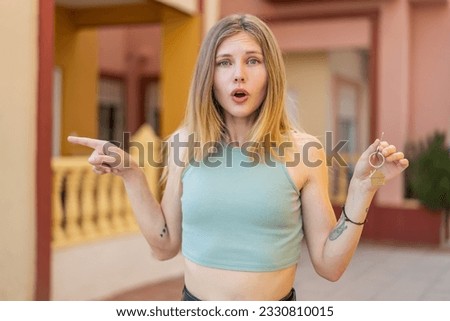Young blonde woman holding home keys at outdoors surprised and pointing side
