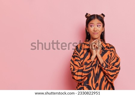 Oriental girl with two buns hairstyle pressing her hands to her chest and looks aside sceptically, posing on pink background, moment of truth concept, copy space