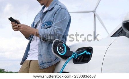 Asian man use phone while electric car recharge energy, showing digital battery status hologram from EV charger from charging station at wind turbine farm. Alternative clean energy utilization.Peruse