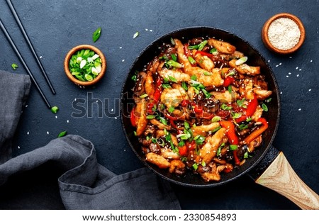 Asian cuisine stir fried chicken, paprika, mushrooms, chives with sesame seeds in frying pan. Black kitchen table background, top view Royalty-Free Stock Photo #2330854893