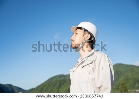 A Japanese male worker wearing a helmet and work clothes stares into the distance against a background of Japanese countryside. Copy space available.