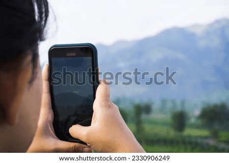 A woman wearing goose down jacket is taking a photo of a mountain and plantation landscape in the morning using her smartphone. Merapi Merbabu Mountainside in Selo Boyolali, Central Java, Indonesia.