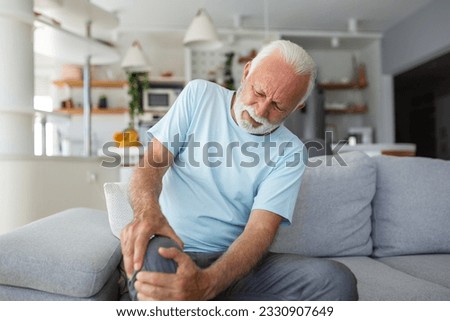 The older man is sitting on the couch at home, has pain in the knee joint, holding his leg, osteoarthritis concept. Royalty-Free Stock Photo #2330907649