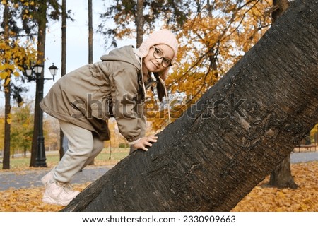 Girl child in glasses, a pink hat and an autumn jacket climbs up a tree in an autumn park. Horizontal photo