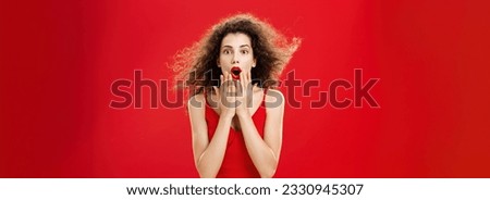 Attractive elegant young woman in red evening dress. with curly hair flicking on wind saying wow being impressed and thrilled covering opened mouth from amazement standing over red background. Royalty-Free Stock Photo #2330945307