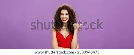 Suggesting you check out. Charming joyful and friendly stylish european woman with curly hairstyle in evening red dress pointing left with thumb and smiling assured and confident showing copy space.