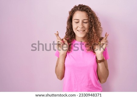 Young caucasian woman standing over pink background gesturing finger crossed smiling with hope and eyes closed. luck and superstitious concept. 