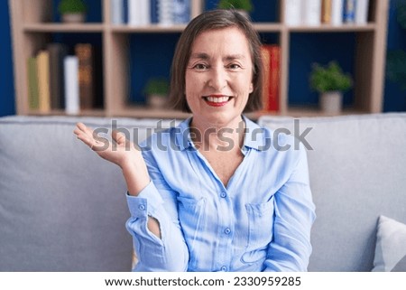 Middle age hispanic woman sitting on the sofa at home smiling cheerful presenting and pointing with palm of hand looking at the camera. 