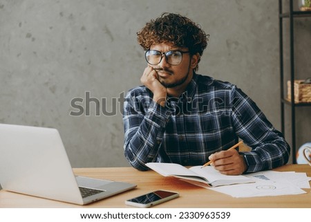 Tired troubled exhausted sad weary employee business Indian man he wearing casual blue checkered shirt looking camera writing in notebook sit working at office desk with laptop pc computer indoors Royalty-Free Stock Photo #2330963539