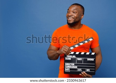 Young fun man of African American ethnicity he wear orange t-shirt hold in hand classic black film making clapperboard look aside isolated on plain dark royal navy blue background. Lifestyle concept