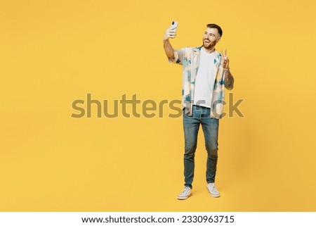 Full body young man he wear blue shirt white t-shirt casual clothes doing selfie shot on mobile cell phone post photo on social network show v-sign isolated on plain yellow background studio portrait