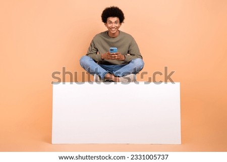 Full length photo of cheerful young man sit empty space podium use smart phone isolated on beige color background