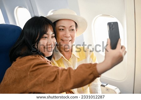 A happy Asian grandmother is taking photos with a smartphone with her granddaughter during the flight for their summer vacation together. Journey, summer trip, airplane, lifestyle